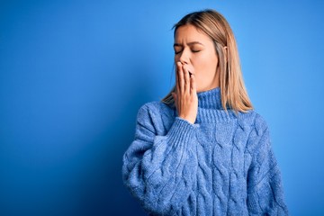 Young beautiful blonde woman wearing winter wool sweater over blue isolated background bored yawning tired covering mouth with hand. Restless and sleepiness.