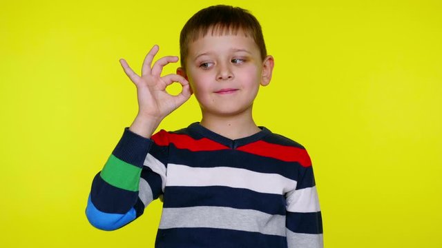 Cheerful little kid boy in a multi-colored sweater raises his hand, shows ok and smiles on a yellow background with copy space. Place for text or product. 4K slowmotion footage