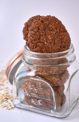 Open lid glass cookie jar with oatmeal cookies inside with oat flakes spread on the white surface table - delicious gluten-free oatmeal cookies with walnuts and cocoa