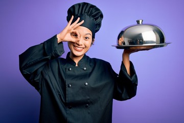 Young beautiful chinese chef woman wearing cooker uniform and hat holding tray with dome with happy face smiling doing ok sign with hand on eye looking through fingers