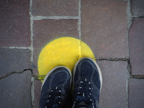 Human Legs And Feet Waiting In Line And Standing Inside Marked Yellow Circles With Social Distancing Measures. Yellow Road Marking And Sidewalk Symbol.