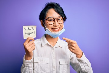Young beautiful asian girl wearing medical mask holding reminder with virus alert message very happy pointing with hand and finger