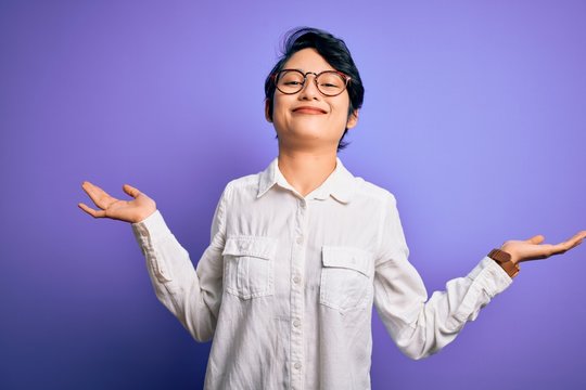 Young beautiful asian girl wearing casual shirt and glasses standing over purple background clueless and confused expression with arms and hands raised. Doubt concept.