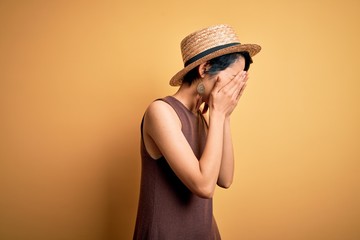 Young beautiful asian girl wearing casual t-shirt and hat over isolated yellow background with sad expression covering face with hands while crying. Depression concept.