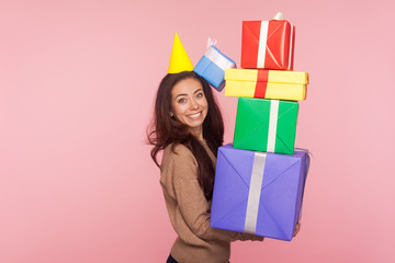 Fototapeta premium Portrait of excited joyful young woman wearing party cone and smiling pleased as holding mount of gift boxes, enjoying many best presents, celebrating birthday. studio shot isolated on pink background