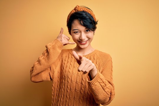 Young beautiful asian girl wearing casual sweater and diadem standing over yellow background smiling doing talking on the telephone gesture and pointing to you. Call me.