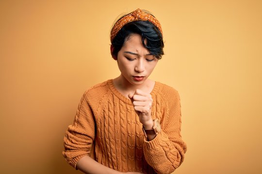 Young beautiful asian girl wearing casual sweater and diadem standing over yellow background feeling unwell and coughing as symptom for cold or bronchitis. Health care concept.