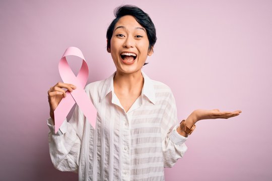 Young beautiful asian girl holding pink cancer ribbon symbol over isolated background very happy and excited, winner expression celebrating victory screaming with big smile and raised hands