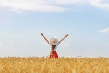 Fototapeta na wymiar Girl in hat stands in middle of wheat field raising his hands to sky