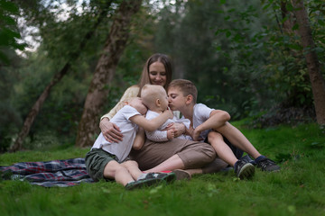 Naklejka na ściany i meble Mother and three children sitting on picnic blanket in park. Happy friendly family. Two brothers hug little sister.