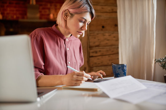 Side view of serious young female outsourced bookeeper providing accounting service for small businesses, sitting at desk with portable computer, holding pen to make notes and using calculator