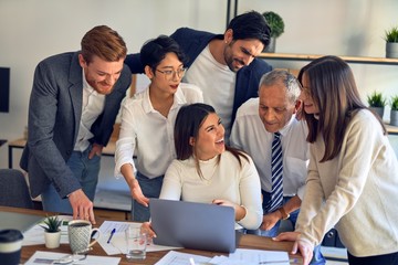 Group of business workers smiling happy and confident. One of them sitting and partners standing around. Working together with smile on face looking at the laptop at the office