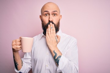 Handsome bald man with beard and tattoo drinking cup of coffee over pink background cover mouth...