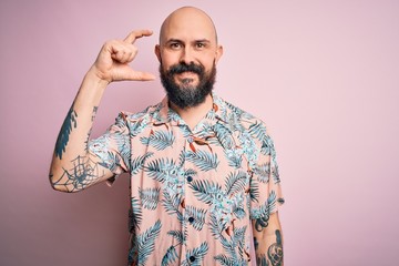 Handsome bald man with beard and tattoo wearing casual floral shirt over pink background smiling and confident gesturing with hand doing small size sign with fingers looking and the camera. Measure