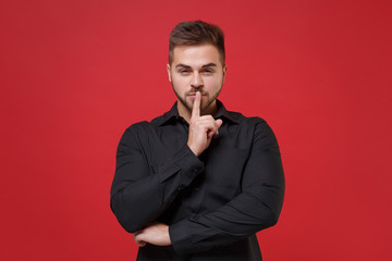 Secret young bearded guy 20s in classic black shirt posing isolated on red background studio portrait. People lifestyle concept. Mock up copy space. Say hush be quiet with finger on lips shhh gesture.