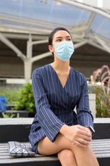 Young Asian businesswoman with mask sitting and thinking in the city