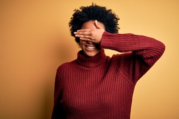 Fototapeta na wymiar Young beautiful African American afro woman with curly hair wearing casual turtleneck sweater smiling and laughing with hand on face covering eyes for surprise. Blind concept.