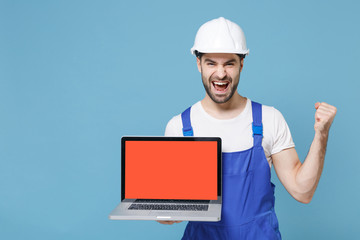 Happy man in coveralls hardhat hold laptop computer with blank empty screen isolated on blue background. Instruments accessories renovation apartment room. Repair home concept. Doing winner gesture.
