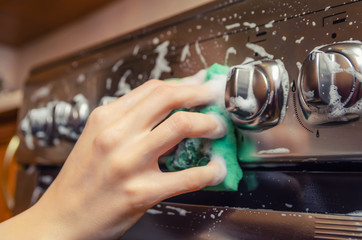 woman's hand close up washes a green sponge with foam on the switch handle of a gray stainless steel gas stove. Cleaning the apartment