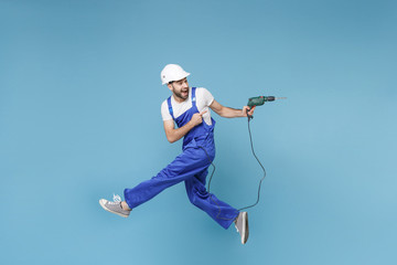 Side view of excited young man in coveralls protective helmet hardhat hold electric drill isolated on blue background. Instruments accessories renovation apartment room. Repair home concept. Jumping.