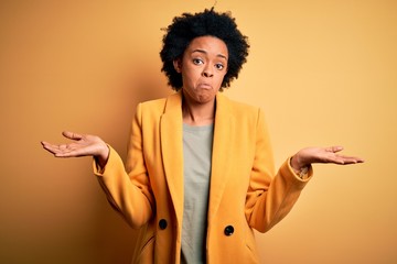 Young beautiful African American afro businesswoman with curly hair wearing yellow jacket clueless...