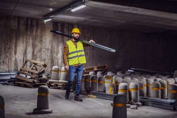 Full length of handsome caucasian bearded worker in vest and helmet on head carrying metal rod on...
