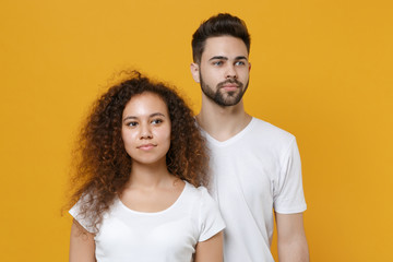 Beautiful young couple friends european guy african american girl in white t-shirts posing isolated on yellow wall background in studio. People lifestyle concept. Mock up copy space. Looking aside.