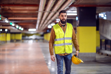 Three quarter length of handsome bearded worker in vest holding his safety helmet and going back to...