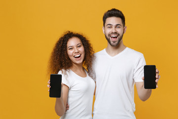 Excited young couple two friends european guy african american girl in white t-shirts posing isolated on yellow background. People lifestyle concept. Hold mobile phone with blank empty screen hugging.