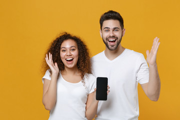 Excited couple two friends european guy african american girl in white t-shirts isolated on yellow background. People lifestyle concept. Hold mobile phone with blank empty screen, spreading hands.