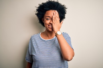 Young beautiful African American afro woman with curly hair wearing striped t-shirt covering one eye with hand, confident smile on face and surprise emotion.