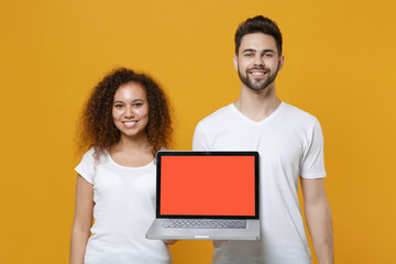 Smiling young couple two friends european guy african american girl in white t-shirts isolated on yellow background studio. People lifestyle concept. Hold laptop pc computer with blank empty screen.