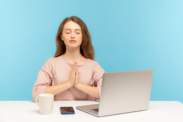 Naklejka premium Break at work, yoga practice. Calm woman employee sitting at workplace with laptop and raising hands in namaste gesture, meditating at home office. indoor studio shot isolated on blue background