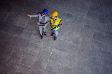 Top view of supervisor with helmet on head in suit standing with construction worker, holding...