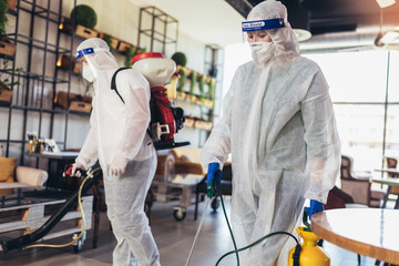 Professional workers in hazmat suits disinfecting indoor of cafe or restaurant, pandemic health...
