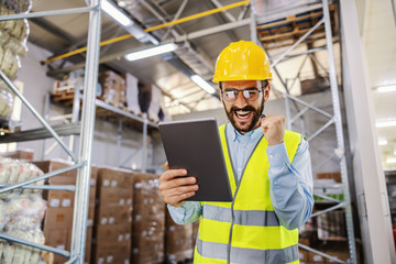 Young smiling supervisor in vest and protective helmet standing in warehouse and using tablet for...