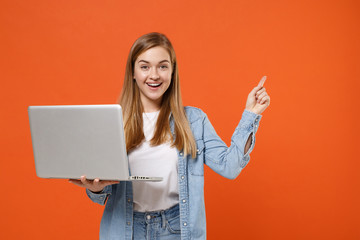 Cheerful young woman girl in casual denim clothes posing isolated on orange background studio portrait. People lifestyle concept. Mock up copy space. Hold laptop pc computer, pointing index finger up.