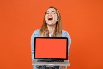 Laughing young woman girl in casual denim clothes posing isolated on orange background studio portrait. People lifestyle concept. Mock up copy space. Hold laptop pc computer with blank empty screen.