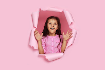 Portrait of cute surprised caucasian little child girl looking with happy face expression at the camera in torn paper pink wall. copy space. banner for discounts and sales