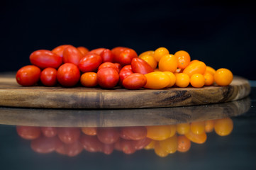 cherry tomatoes, red and yellow on a wooden cutting board with a black background and a mirrored...