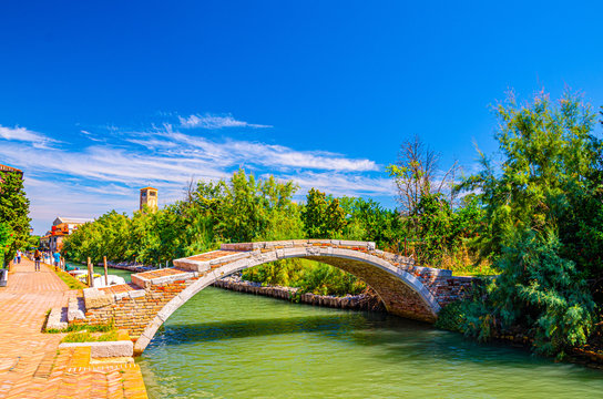 Stone Devil bridge across water canal on Torcello island, embankment promenade along water canal, green trees, tower and blue sky background. Venetian Lagoon, Veneto Region, Northern Italy.