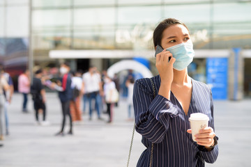 Young Asian businesswoman with mask talking on phone while having coffee on the go as the new...