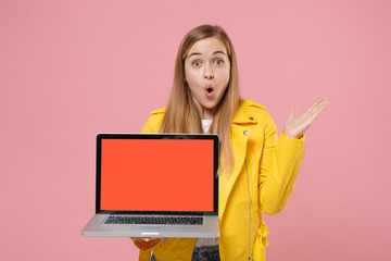 Shocked woman in yellow leather jacket isolated on pastel pink background. People lifestyle concept. Mock up copy space. Hold laptop pc computer with blank empty screen, spreading pointing hand aside.