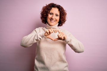 Middle age beautiful curly hair woman wearing casual turtleneck sweater over pink background smiling in love doing heart symbol shape with hands. Romantic concept.