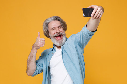 Cheerful elderly gray-haired mustache bearded man in casual blue shirt isolated on yellow background. People lifestyle concept. Mock up copy space. Doing selfie shot on mobile phone showing thumb up.