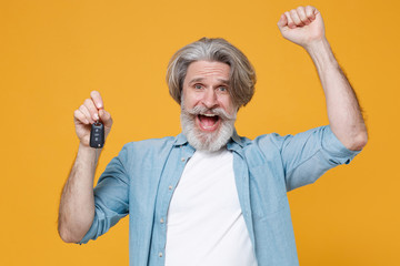 Joyful elderly gray-haired mustache bearded man in casual blue shirt posing isolated on yellow wall background studio. People lifestyle concept. Mock up copy space. Hold car keys doing winner gesture.