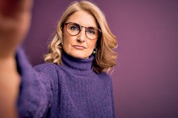 Middle age beautiful blonde woman wearing sweater and glasses make selfie by the camera with a confident expression on smart face thinking serious