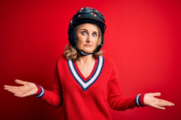 Middle age beautiful blonde motorcyclist woman wearing moto helmet over red background clueless and confused expression with arms and hands raised. Doubt concept.