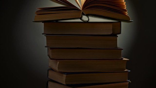 Elevating past a stack of books to see an open book on top of them. Yellowed pages of the old open book waving in wind. Crane shot, UHD