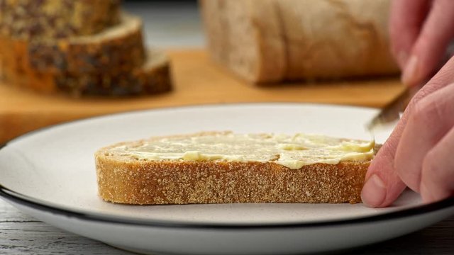 Female hands spreading butter on a slice of bread using knife. Two types of bread blurred in background. Slider shot, 4K